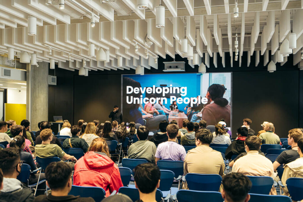 Students attending a teen program presentation with a large screen reading “Developing Unicorn People” in a modern lecture space.